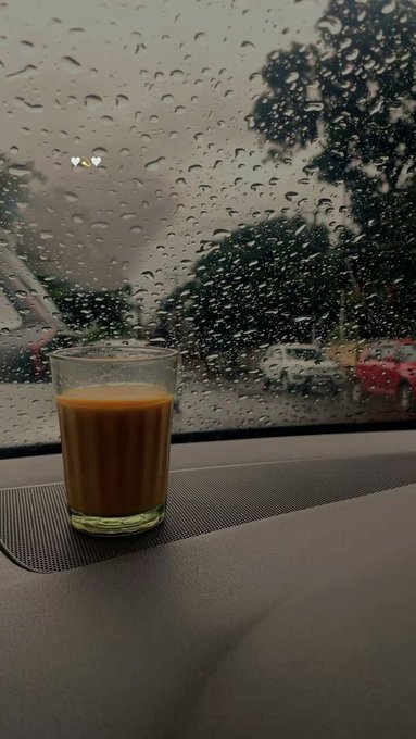 A glass of an orange-colored beverage, possibly juice or coffee, sits on the dashboard of a car with raindrops on the windshield and a blurred street scene visible outside.