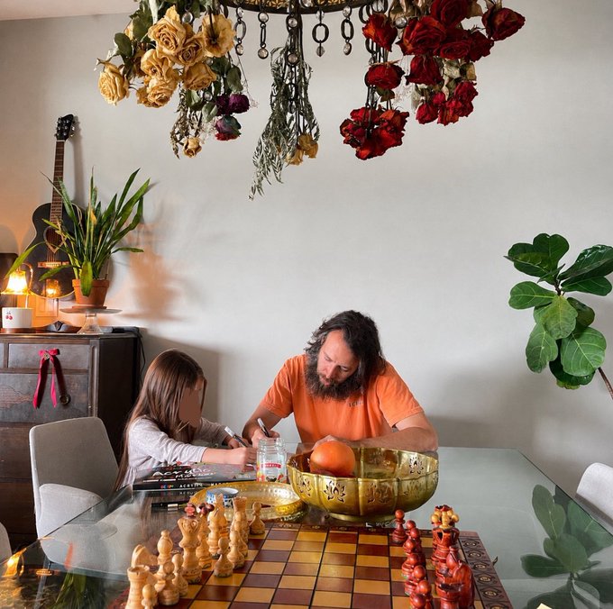 A photograph of a man and a child playing chess at a glass table, with hanging dried flowers and a guitar visible in the background.