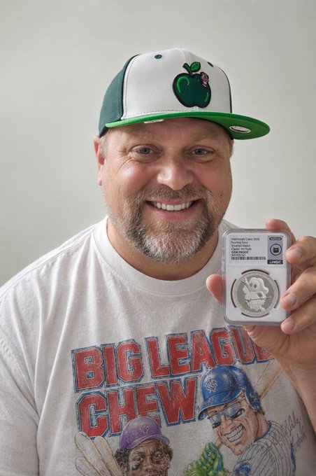 A smiling bearded man wearing a green and white 'Boston Celtics' apple logo cap and a 'Big League Chew' t-shirt proudly holds up an NGC-graded Pikachu coin in a protective slab.