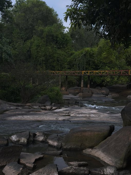 A scenic view of a rocky stream flowing through a forested area with a yellow metal truss bridge crossing the water in the background.