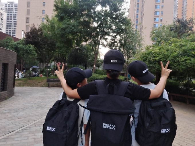 Three young people from behind, wearing matching black caps and black 'OKX' branded backpacks, posing with peace signs in a park area with apartment buildings in the background.