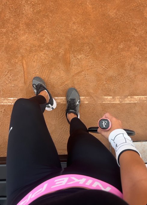 First-person view looking down at someone's feet on a clay tennis court, wearing grey Nike shoes, black leggings, and holding a pink and black Nike paddle while wearing a white wristband.