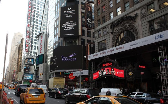 A street view of Times Square in New York City featuring bright billboards, including advertisements for 'PF Newswire', 'Mubi', 'Pussy Manager', 'Hard Rock Hotel & Casino', and 'Lids', with yellow taxis driving on the street below.