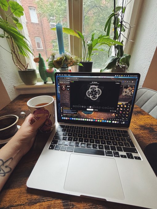A first-person view of someone holding a mug of tea while sitting at a wooden table with an open laptop displaying a retro video game, surrounded by potted plants on a windowsill.