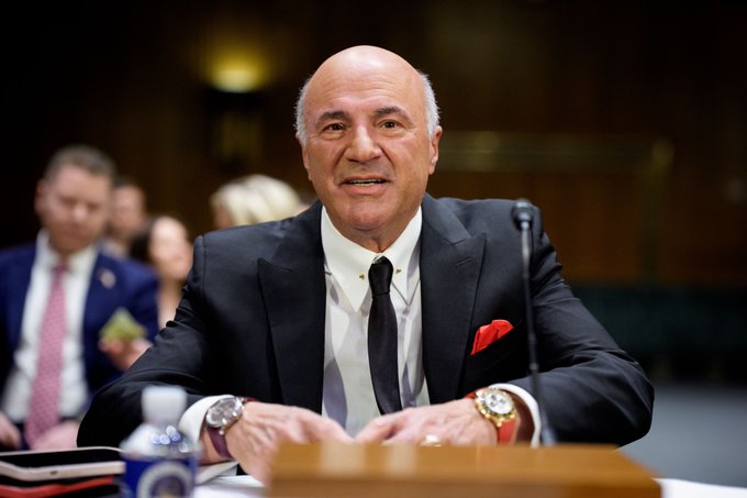Kevin O'Leary sits at a table during what appears to be a congressional hearing, wearing a dark suit with a red pocket square and multiple watches, as he reportedly discusses the CLARITY Act's uncertain prospects and concerns about Iran tensions affecting legislative priorities.