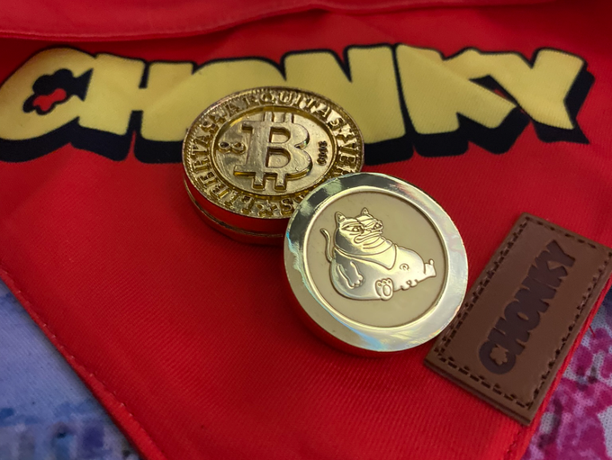 Two metallic coins rest on a red fabric with the 'CHUNKY' logo, including a Bitcoin-style coin and one featuring a chubby cat, alongside a leather tag stamped with 'CHUNKY'.