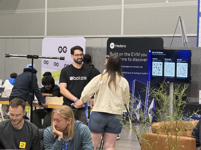 People at what appears to be a conference or tech event, with a man wearing a 'bolos' branded shirt standing near a 'Hedera' booth and digital screens displaying QR codes.