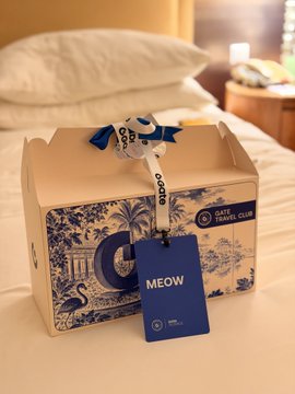 A decorative blue and white gift box labeled "GATE TRAVEL CLUB" sits on a hotel bed with a personalized "MEOW" tag and ribbon, presented to Gate Travel Club members as a thoughtful welcome amenity during their Bali trip.