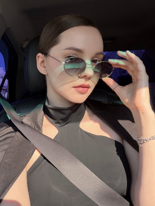 A woman sits in a car wearing round sunglasses and a black top, holding the sunglasses with one hand while wearing a seatbelt.