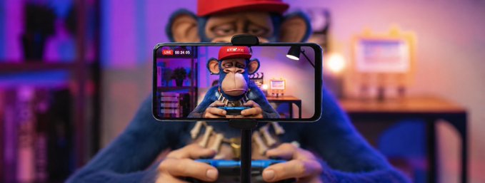 A person holds a smartphone displaying a monkey mascot in a red cap, while the same character appears as a 3D figure behind the phone in a purple-lit room.
