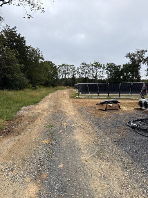 A dirt and gravel pathway leads through a rural area past a solar panel array, with trees and an overcast sky in the background.