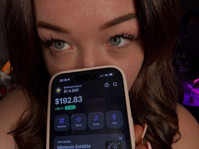 Selfie of a woman holding up a phone displaying a crypto portfolio showing '$1 A DAY' by @allergictogueuc with $192.83 in holdings including Memecoin $ANSEM.