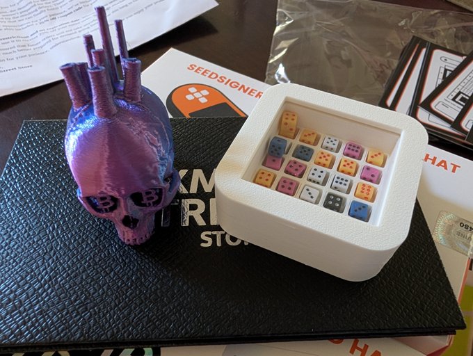 A purple 3D-printed Bitcoin-shaped object sits on a black notebook next to a SeedSigner hardware wallet box containing colorful dice.