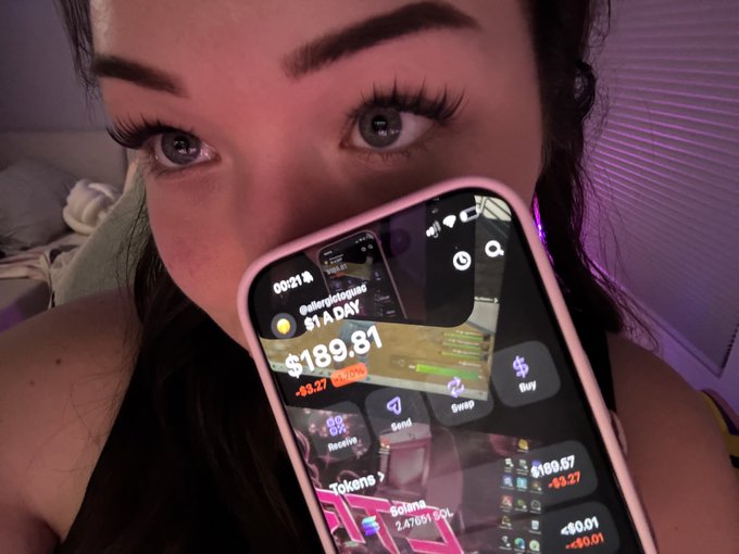 A close-up selfie of a young woman holding a smartphone displaying a crypto portfolio with $189.81 and $169.57 values, with her eyes visible above the pink phone case.