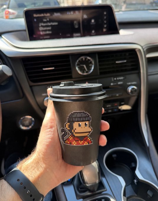 A hand holding a black coffee cup featuring pixel-art character artwork, held up inside a car (appears to be a Lexus) with the dashboard and infotainment screen visible.