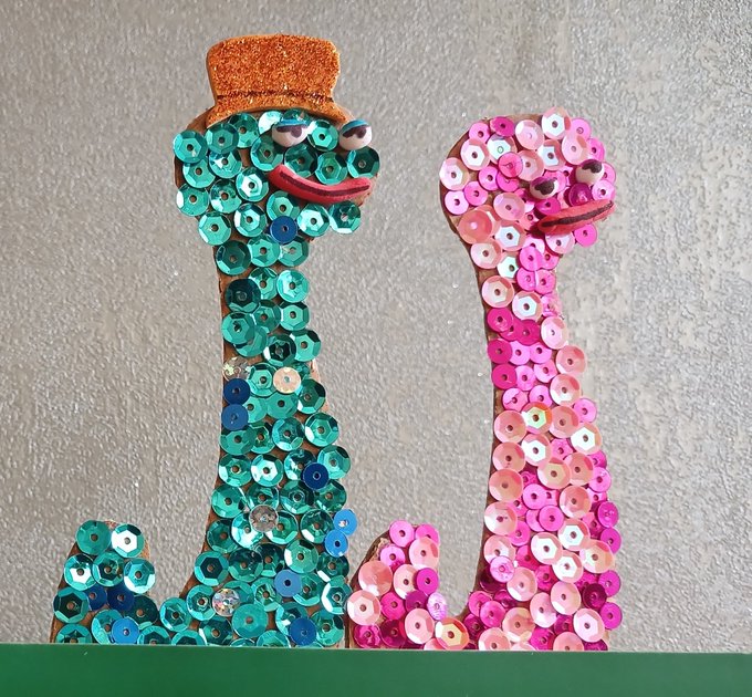 Two handcrafted snake figures decorated with sequins—one in teal/blue wearing an orange top hat and one in pink with googly eyes—displayed on a green surface against a gray wall.