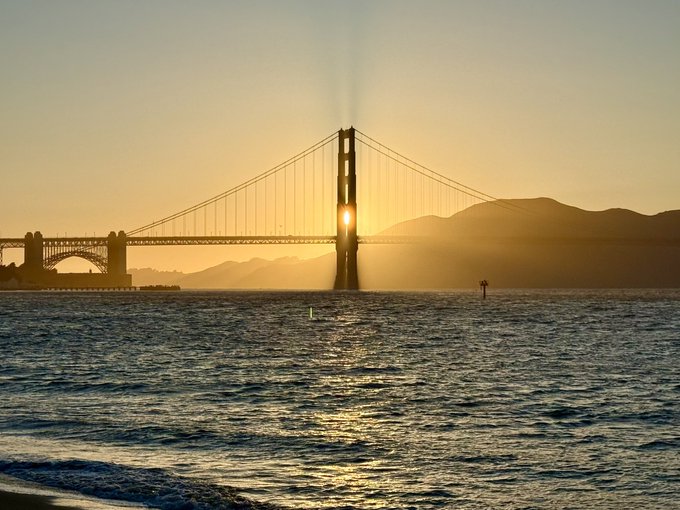 The image features a beautiful sunset over the ocean, with the Golden Gate Bridge visible in the background. The bridge spans across the water, creating a stunning backdrop for the scene. The sun's rays are shining down on the water, causing it to sparkle and create a warm glow that illuminates the entire scene. In the foreground of the image, there is a person standing near the water, possibly enjoying the view or taking a photograph of the sunset.