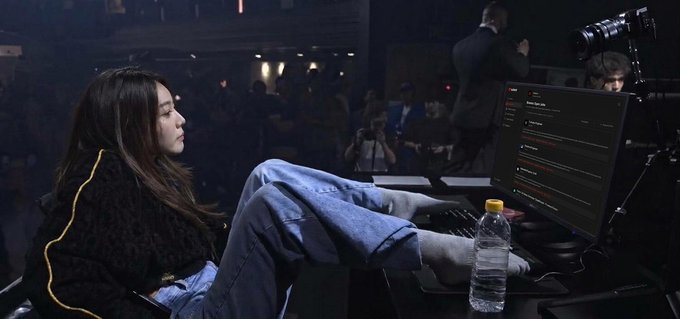 A young woman with long dark hair sits at an esports gaming setup in a dimly lit arena, typing at a keyboard with feet propped up, surrounded by photographers and an audience in the background.