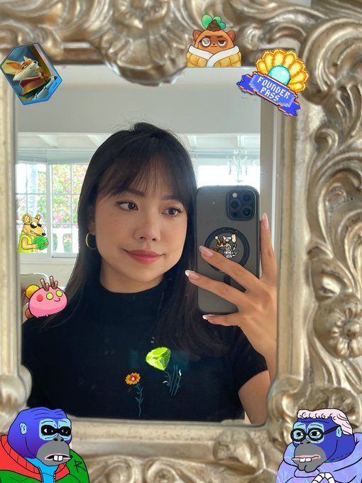 A selfie of a young woman with dark hair in a black shirt taking a mirror photo with her phone, framed by an ornate gold mirror and decorated with various cartoon stickers including monkeys and 'Founder Pass' badges.