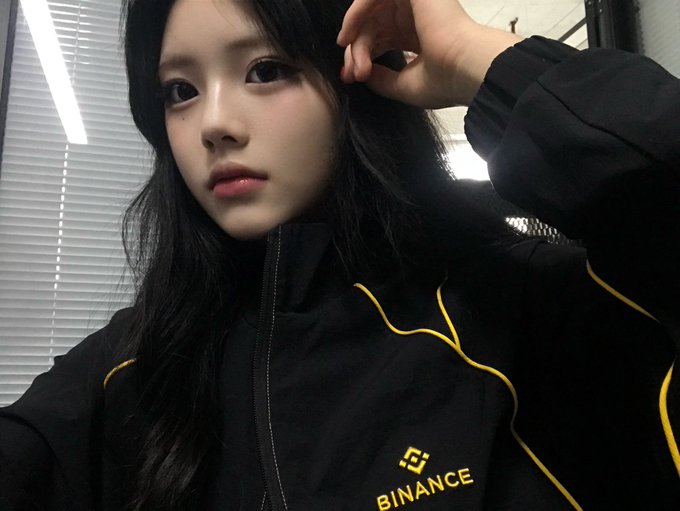 A young woman poses for a selfie wearing a black jacket with yellow piping and a 'BINANCE' logo.