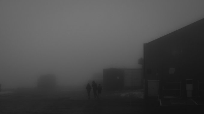 A foggy, low-visibility scene shows several silhouetted figures walking near a dark building, with another structure barely visible in the misty background.