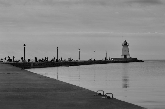 A black and white photograph of a pier extending into calm water, ending with a small lighthouse on the right under a cloudy sky.
