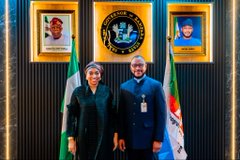 Two people in dark suits standing in front of a Nigerian government flag and official seal, with framed portraits on the wall behind them.