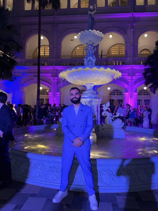 A man with a ponytail poses in a light blue double-breasted suit in front of an illuminated fountain at a nighttime event, surrounded by vibrant purple and blue lighting that enhances the elegant, sophisticated atmosphere suggested by "And we're only getting started."