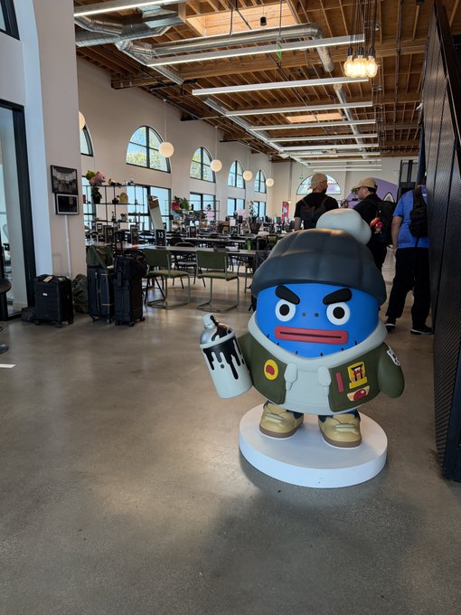 A cartoonish blue Azuki mascot statue in streetwear holds a spray paint can inside the brand's LA headquarters, where visitors browse merchandise and inspect luggage in the industrial-chic space.