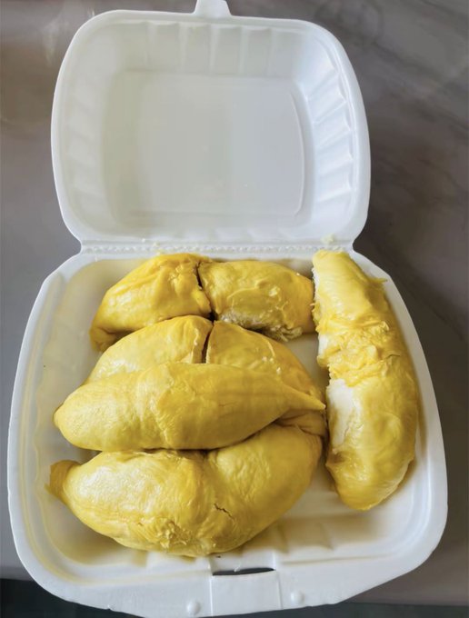 A styrofoam takeout container is open, displaying several pieces of fresh durian fruit with their characteristic bright yellow, creamy flesh visible inside the white compartments.