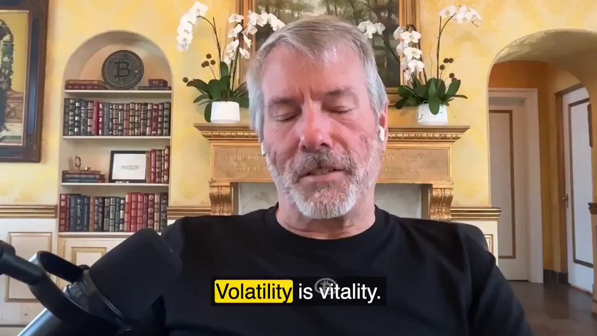 The image features an older man sitting comfortably in a chair while wearing a black shirt. The background includes a fireplace and several bookshelves filled with various books. There are also potted plants placed around the room, adding a touch of greenery to space. The text on the image reads "Volatility is Satoshi's gift to the faithful." This quote is attributed to Saylor Kranke, an expert in Bitcoin and cryptocurrency markets.