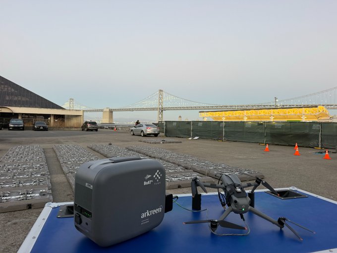 A DJI drone and an ar.green portable power station sit on a blue surface with a large suspension bridge visible in the background across a parking area.