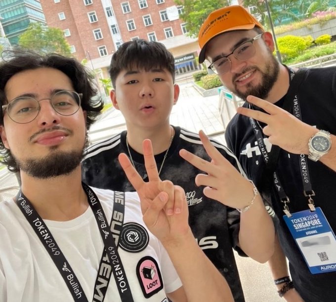 Three young men posing for a selfie at what appears to be the Token2049 Singapore conference, making peace signs and wearing event lanyards.