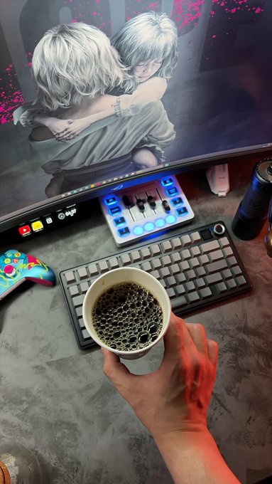 A gaming desk setup showing a person holding a cup of coffee, a keyboard, an audio mixer, and a curved monitor displaying anime art.