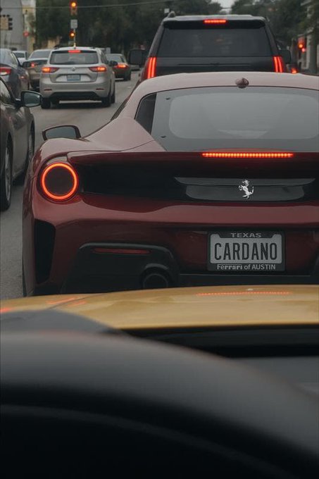 The rear view of a red Ferrari sports car at a traffic stop, featuring a Texas license plate that reads 'CARDANO' and a dealer tag stating 'Ferrari of Austin'.