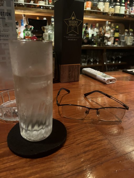 The image shows a table with glasses, a remote control, and a glass of water sitting on top of it. The scene appears to be set at a bar or restaurant where people can enjoy drinks and snacks while watching television. There are multiple bottles visible in the background, suggesting that this is a popular spot for patrons to relax and socialize.