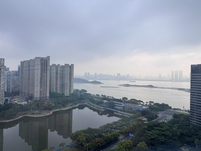A hazy cityscape view overlooks a large body of water, with tall residential buildings lining the left side and a park area in the foreground.