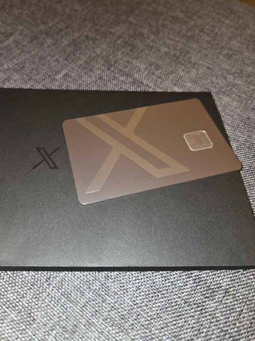 A dark, metallic-looking card with a large embossed 'X' design and a chip rests on top of a black box featuring the XRP cryptocurrency logo.