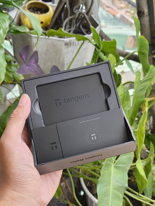 A hand holding an open Tangem Wallet box containing the black card-based hardware wallet, displayed against a backdrop of green plants.