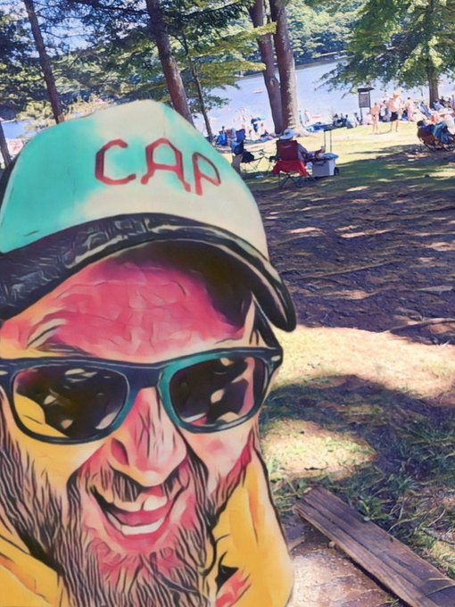 A stylized, heavily filtered photo of a smiling man with a beard and sunglasses wearing a cap with 'CAR' written on it, taken at a park.