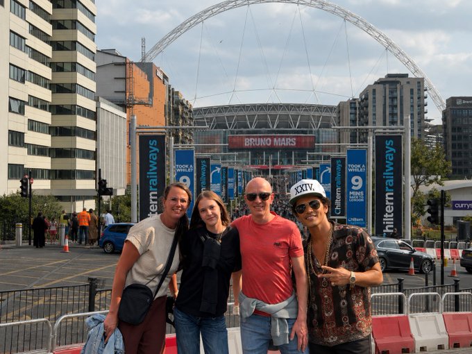 A group of four people posing for a photo outside Wembley Stadium in London, with signage for a Bruno Mars concert and Chiltern Railways visible in the background.