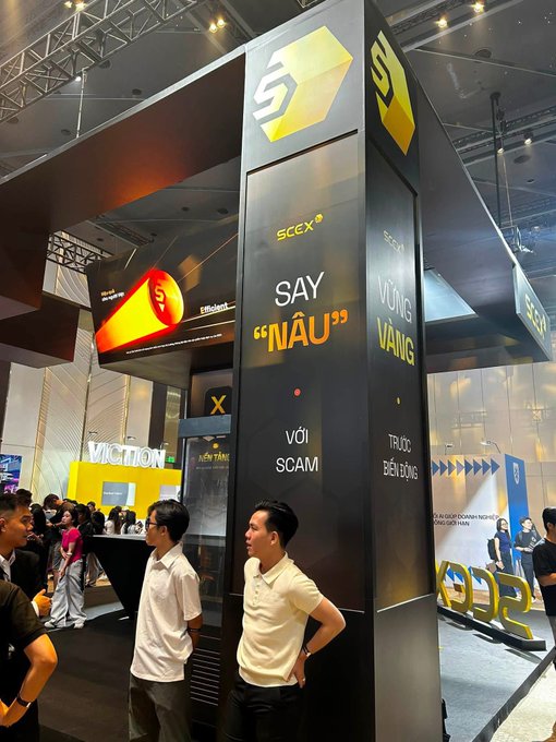 A convention booth for 'SC5X' featuring a large pillar with the text 'SAY "NÂU" VỚI SCAM' and 'TRỤC ĐIỀU BẤT ĐỘNG' surrounded by attendees.