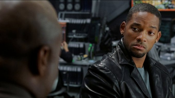 Will Smith, wearing a black leather jacket, looks intently at another man whose back is to the camera, with what appears to be an electronics store or shop visible in the background.
