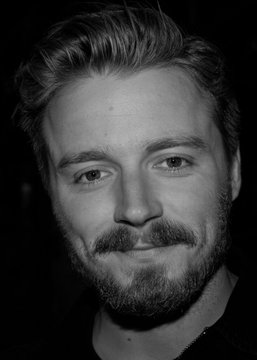 A black-and-white close-up portrait of a bearded man with tousled hair, looking directly at the camera with a subtle smile, accompanying a post about Jack Lowden being the current favorite to be cast as the next James Bond according to Polymarket predictions.