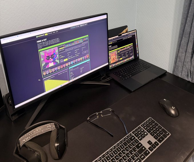 A desk setup featuring a large monitor displaying a gaming website, a laptop, a keyboard, mouse, and headphones on a dark surface.