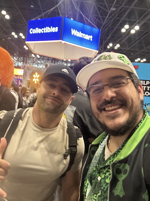 A selfie of two smiling men at what appears to be a convention, standing under a blue illuminated sign reading "Collectibles Walmart."