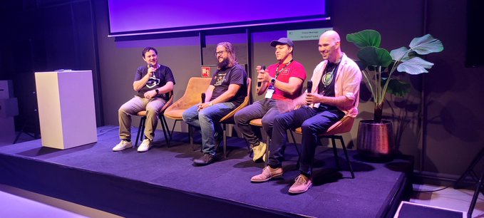 Four men sit on chairs on a stage during a panel discussion, with a potted plant visible in the background.