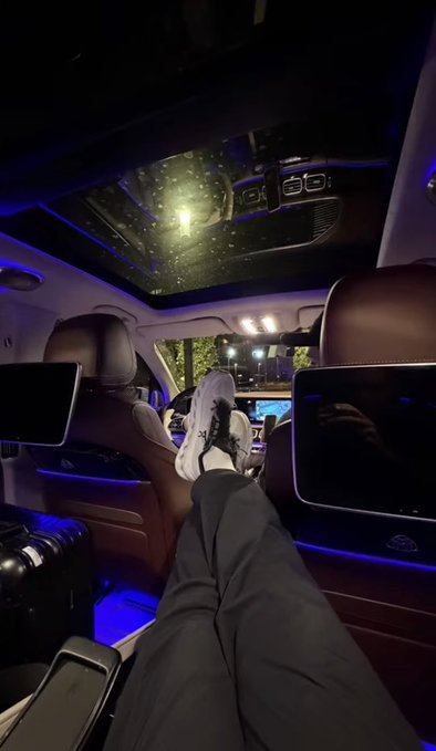 First-person view inside a luxury vehicle at night, showing feet propped up with blue ambient lighting and a starry sky visible through the panoramic sunroof.