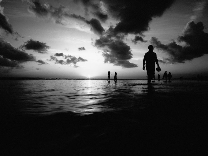 A black-and-white silhouette photograph of people walking along a beach at sunset, with the sun low on the horizon reflecting across the water under dramatic clouds.