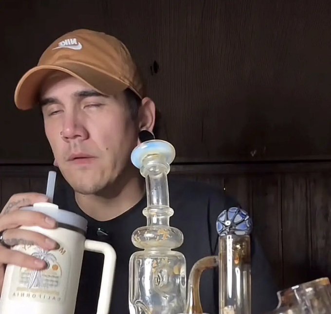 A close-up photograph of a man wearing a tan Nike cap and dark shirt, eyes closed, holding a 'Palm Springs California' branded tumbler with a straw, with glass smoking paraphernalia visible in the foreground.